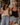 Two women standing outdoors at a cafe, smiling and laughing while holding iced drinks. The woman on the left wears a cream lace camisole and light-wash wide-leg jeans, and the woman on the right wears a black camisole and light-wash denim shorts.